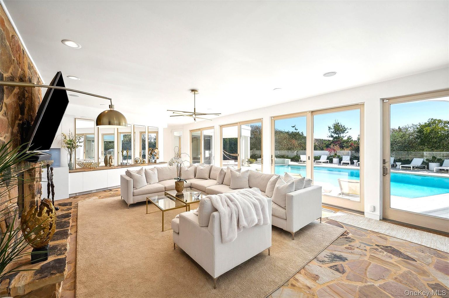 This is a bright and airy living room featuring a large sectional sofa, a glass coffee table, and floor-to-ceiling windows that offer a view of the pool area. The room is decorated in neutral tones, creating a serene and inviting atmosphere. The stone accent wall adds a touch of rustic charm to the modern design.