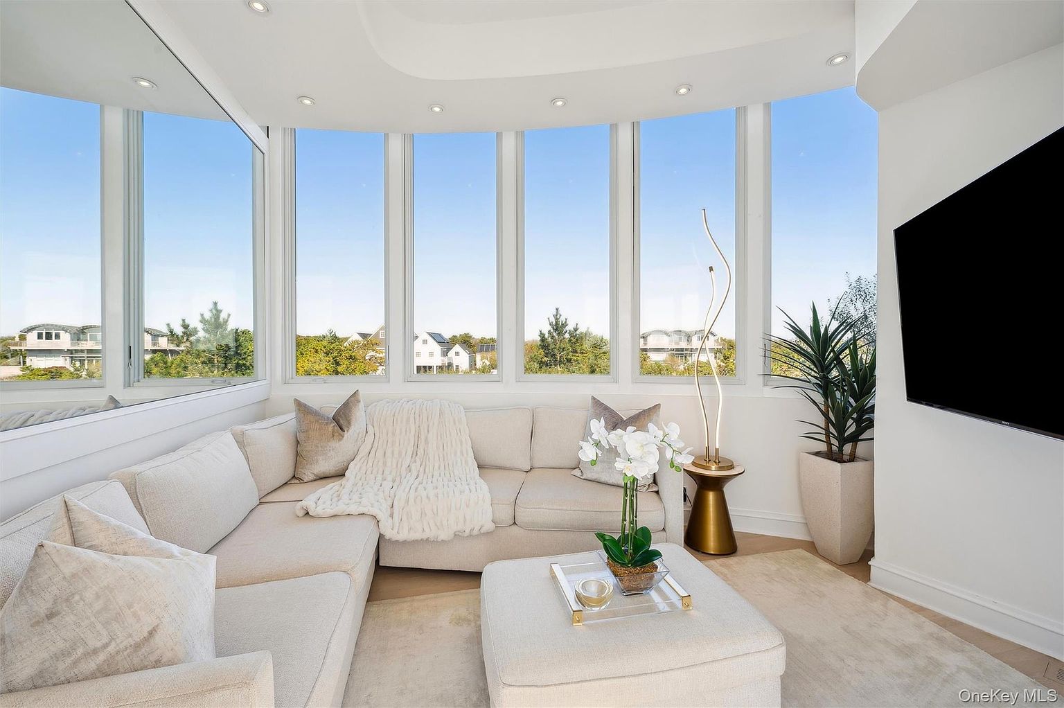 This is an interior shot of a living room featuring a large, comfortable-looking sectional sofa with neutral-toned cushions and a throw blanket. The room is well-lit by natural light streaming through large windows that offer a view of the outdoors. A modern floor lamp and a potted plant add to the room's ambiance, while a television is mounted on the wall.