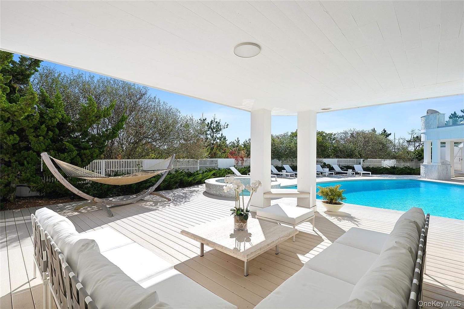 This image showcases a luxurious outdoor patio area with a covered deck, featuring comfortable white sofas and a hammock. The space overlooks a pristine swimming pool and spa, surrounded by lush greenery and a white fence. The overall impression is one of relaxation and upscale outdoor living.