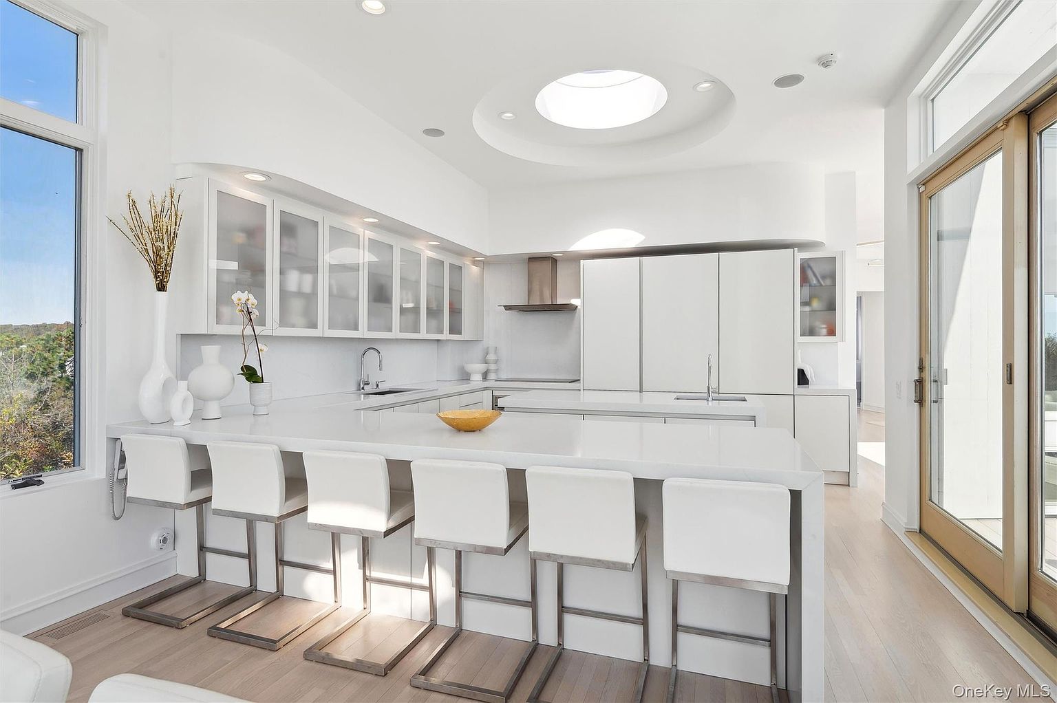 This is a bright, modern kitchen featuring white cabinetry with frosted glass fronts, a large island with seating, and a skylight. The countertops are a clean white, and the overall design is minimalist and airy. The kitchen also features a stainless steel range hood and a large window providing natural light.