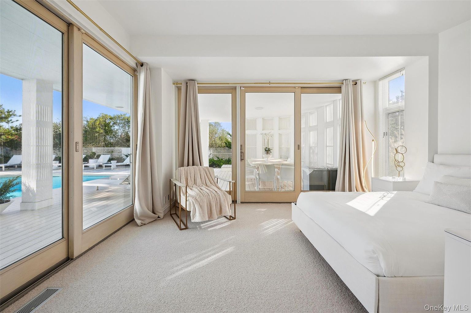 This is a bright and airy primary bedroom featuring large sliding glass doors that lead to an outdoor area with a pool. The room is decorated in a minimalist style with white walls, light carpet, and neutral-toned curtains. A comfortable chair with a throw blanket sits near the doors, and a large bed is positioned on the right side of the room.