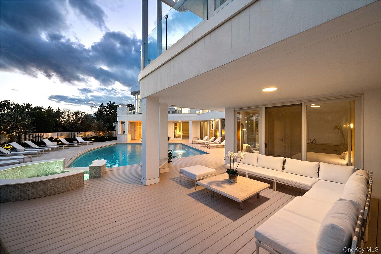 This image showcases a luxurious outdoor patio area with a modern design. The patio features a large white sectional sofa, a coffee table, and a wooden deck. A swimming pool is visible in the background, along with lounge chairs, creating an inviting and relaxing atmosphere.