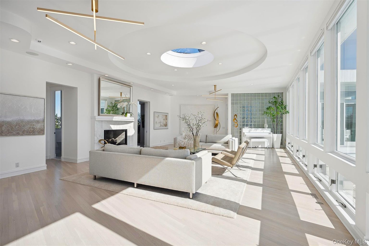 This is a bright and spacious living room featuring a modern design with white walls, light wood floors, and large windows that provide ample natural light. The room includes multiple seating areas with light-colored sofas and chairs, a fireplace with a mirror above, and unique lighting fixtures. A skylight adds to the airy atmosphere, creating an inviting and luxurious space.