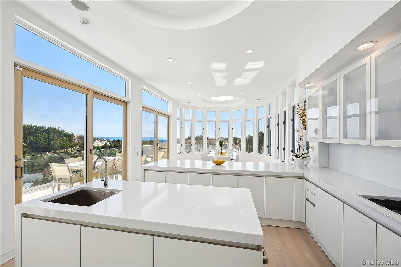 This is a bright, modern kitchen with sleek white cabinetry and countertops. Large windows offer stunning views of the ocean and outdoor patio area, creating a seamless indoor-outdoor living experience. The kitchen features a large island with a sink and ample counter space, perfect for cooking and entertaining.