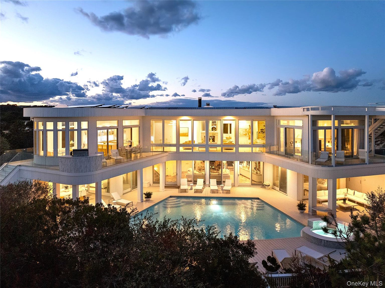 This is an aerial rear view of a modern, luxurious two-story house at dusk. The house features large windows, multiple balconies, and a central courtyard with a swimming pool and hot tub. The lighting inside the house creates a warm and inviting atmosphere, highlighting the architectural design and outdoor amenities.