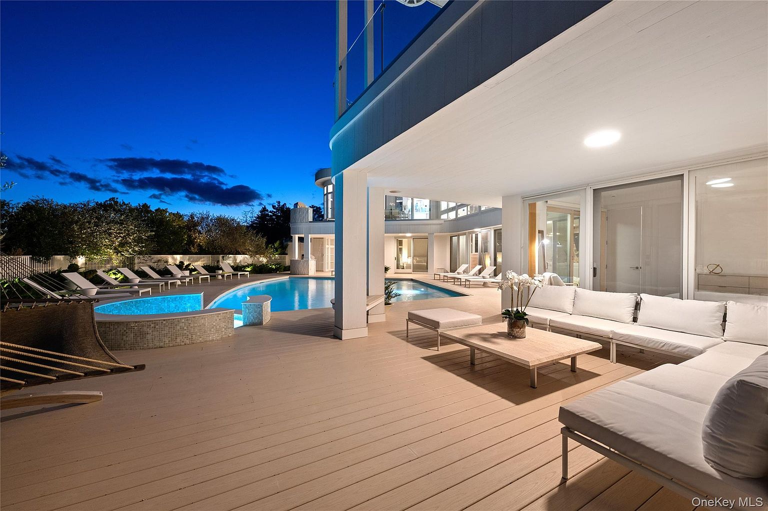 This image showcases a luxurious outdoor patio and deck area, complete with a swimming pool and lounge seating. The deck features modern furniture, including a large sectional sofa and coffee table, creating an inviting space for relaxation and entertainment. The pool is illuminated, adding to the ambiance of the outdoor living area.