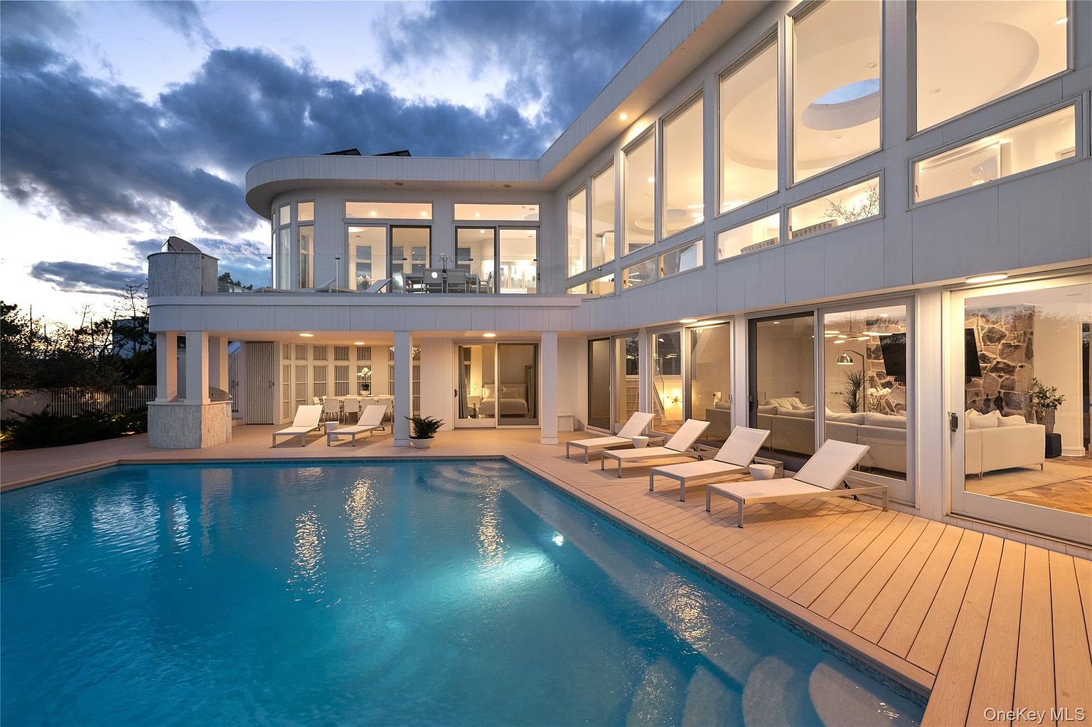 This image showcases the rear exterior of a modern luxury home, emphasizing the pool and outdoor living space. The pool is a prominent feature, surrounded by a wooden deck with lounge chairs. Large glass doors and windows provide seamless indoor-outdoor flow, revealing glimpses of the interior living spaces.