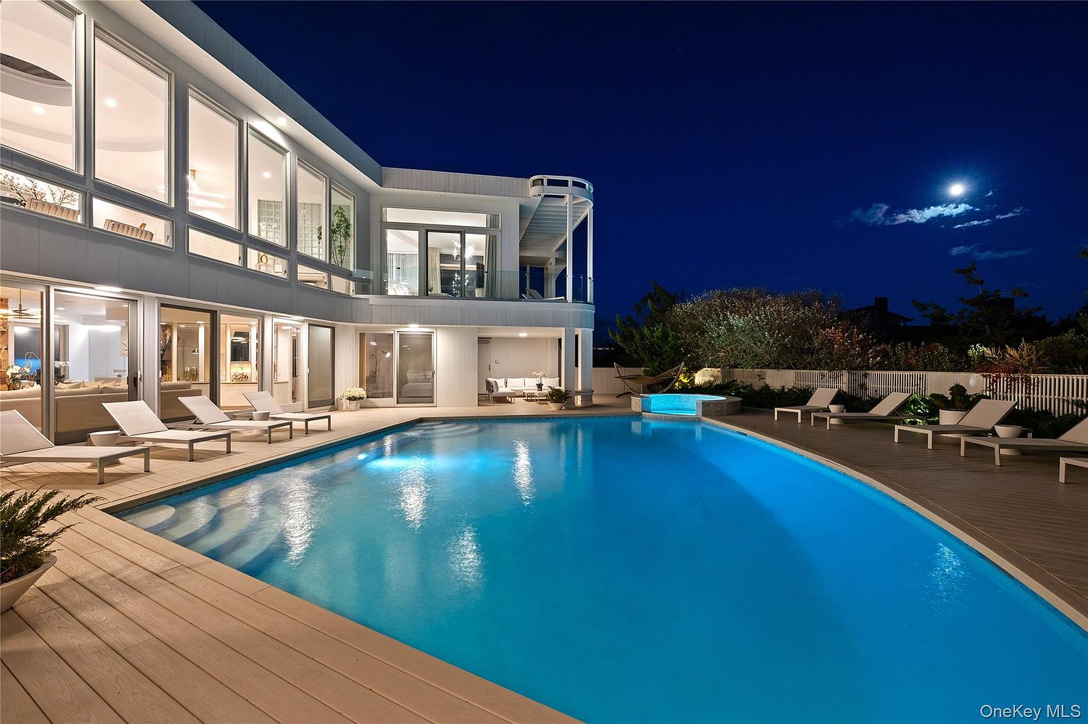This image showcases a luxurious backyard pool and spa area at night. The pool's turquoise water reflects the ambient lighting, and the surrounding deck is furnished with lounge chairs. The modern architecture of the house is visible in the background, with large windows offering glimpses into the interior. The moon is visible in the night sky.