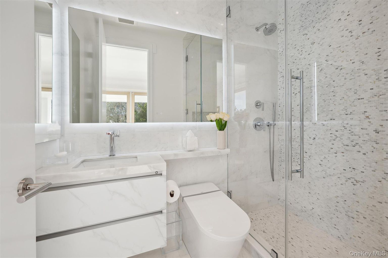This is a modern primary bathroom featuring a sleek vanity with a marble countertop and integrated sink. A large, illuminated mirror hangs above the vanity, reflecting the natural light from a nearby window. The shower is enclosed with glass and features mosaic tile walls, creating a luxurious and spa-like atmosphere.