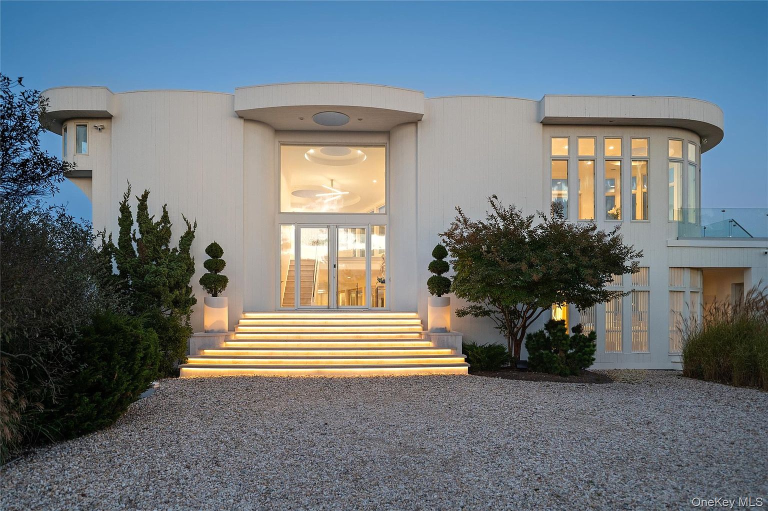 This is a front exterior view of a modern, luxurious home. The house features a white facade, a grand entrance with illuminated steps, and large windows. The property is surrounded by well-maintained landscaping and a gravel driveway, creating an inviting and upscale impression.