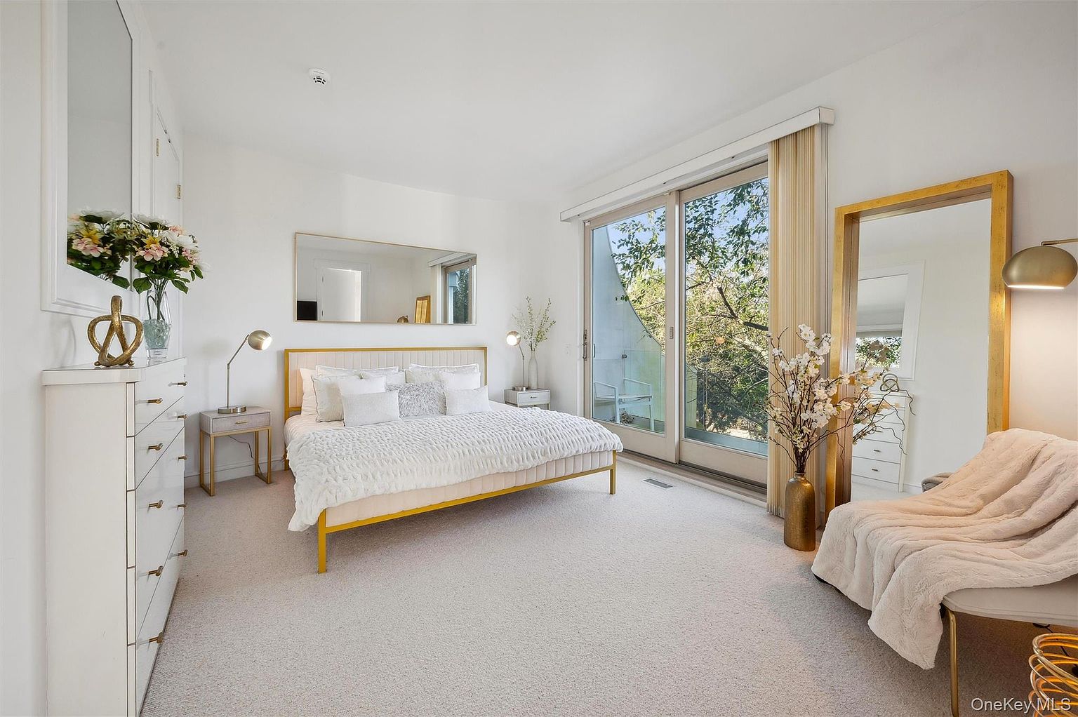 This is a bright and airy primary bedroom featuring a gold-framed bed with white bedding, complemented by a large window with natural light. The room includes a full-length mirror and a comfortable chair with a throw, creating a relaxing and luxurious atmosphere. The neutral color palette and minimalist decor enhance the sense of spaciousness.