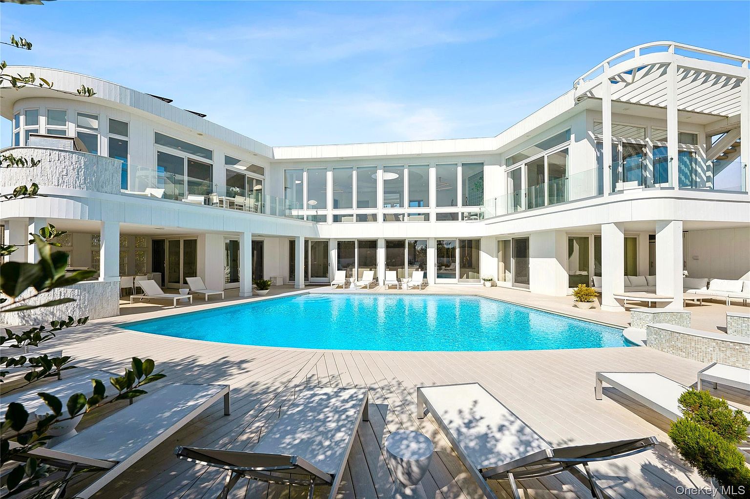 This image showcases a luxurious backyard featuring a large, inviting pool surrounded by a spacious deck with lounge chairs. The modern, white architectural design of the house creates a stunning backdrop, with large windows offering views of the interior. The scene exudes relaxation and upscale living, perfect for entertaining or enjoying a private oasis.