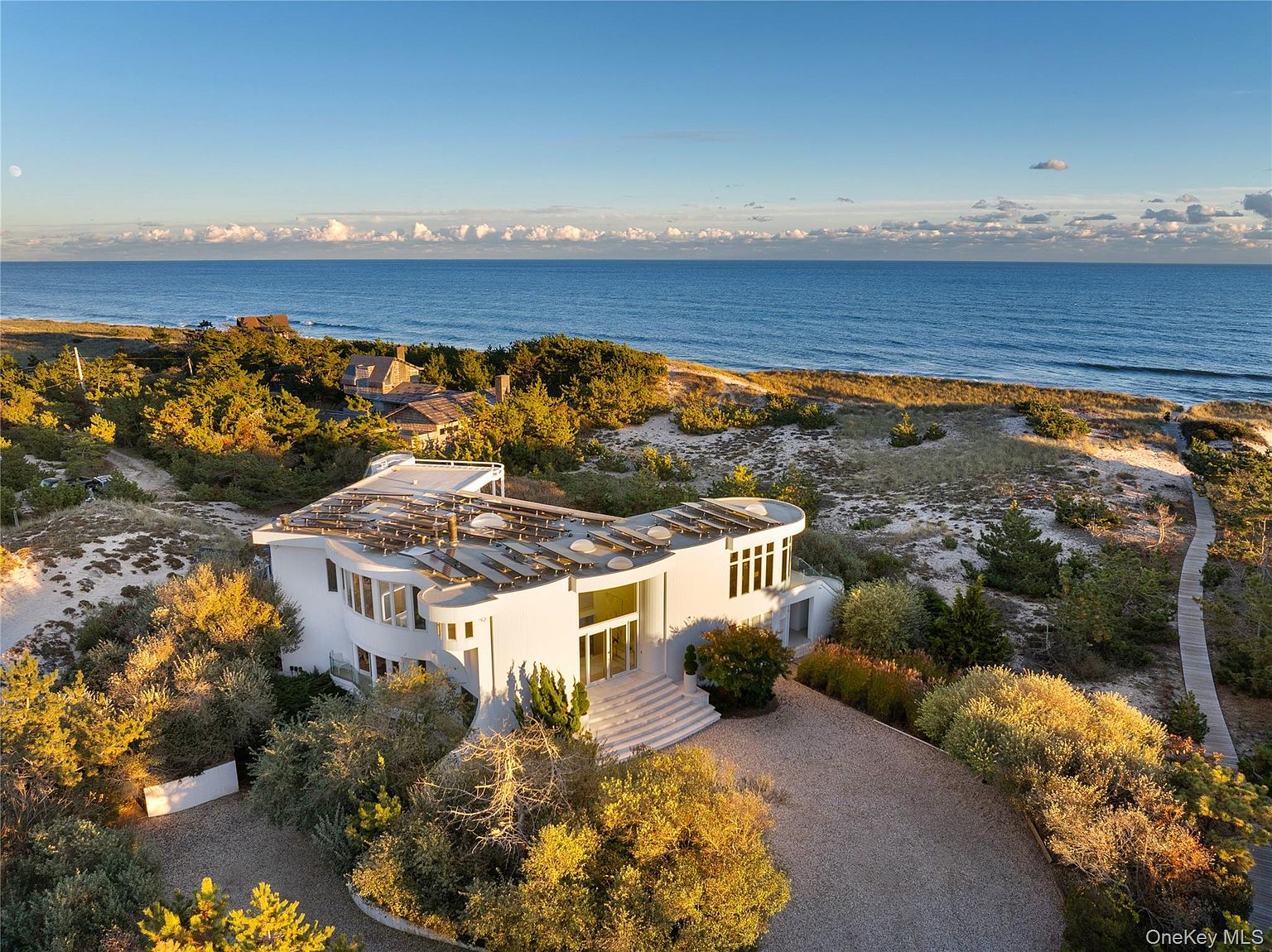 This aerial shot showcases a modern, white architectural masterpiece nestled amidst lush greenery and sandy dunes, with the vast ocean stretching out to the horizon. The unique roof design and expansive windows highlight the home's contemporary style, while the surrounding landscape offers privacy and a connection to nature. A winding driveway leads to the entrance, suggesting a secluded and luxurious property.