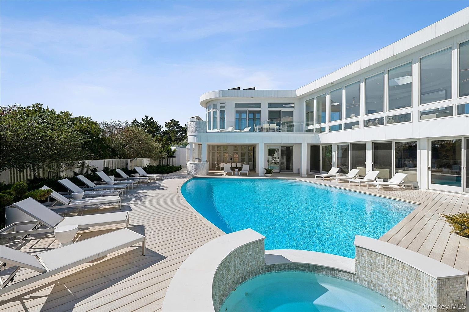 This image showcases a luxurious backyard featuring a large, inviting pool and a separate spa area. The pool is surrounded by a wooden deck with multiple lounge chairs, perfect for relaxation. The modern, white architectural style of the house is visible in the background, with large windows overlooking the pool area, creating a seamless indoor-outdoor living experience.