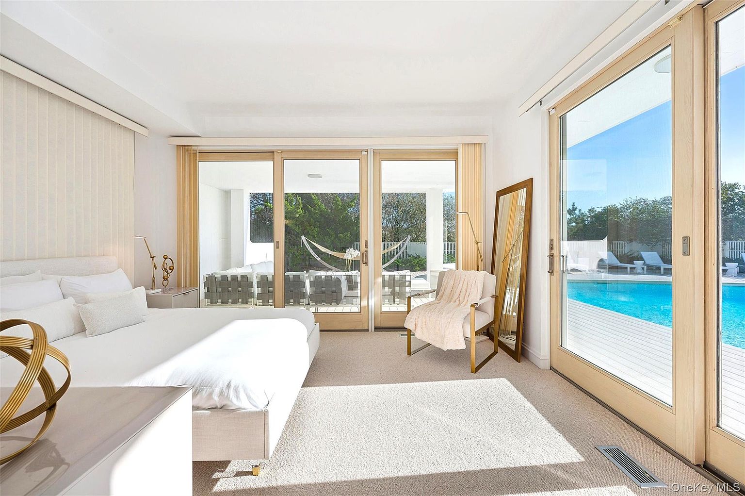 This is a bright and airy primary bedroom with a modern aesthetic. The room features a large bed with white linens, a sitting area with a stylish chair, and sliding glass doors that lead to an outdoor pool area. The neutral color palette and abundance of natural light create a serene and luxurious atmosphere.