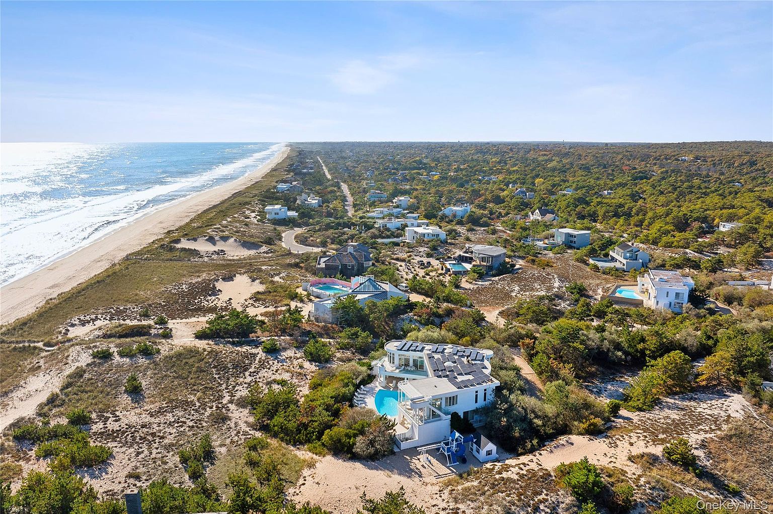 This aerial view showcases a modern beachfront property with a unique architectural design, featuring a curved roof and solar panels. A private pool and outdoor recreational area are visible, nestled amidst lush greenery and sandy dunes. The property offers stunning ocean views and direct beach access, making it a luxurious coastal retreat.