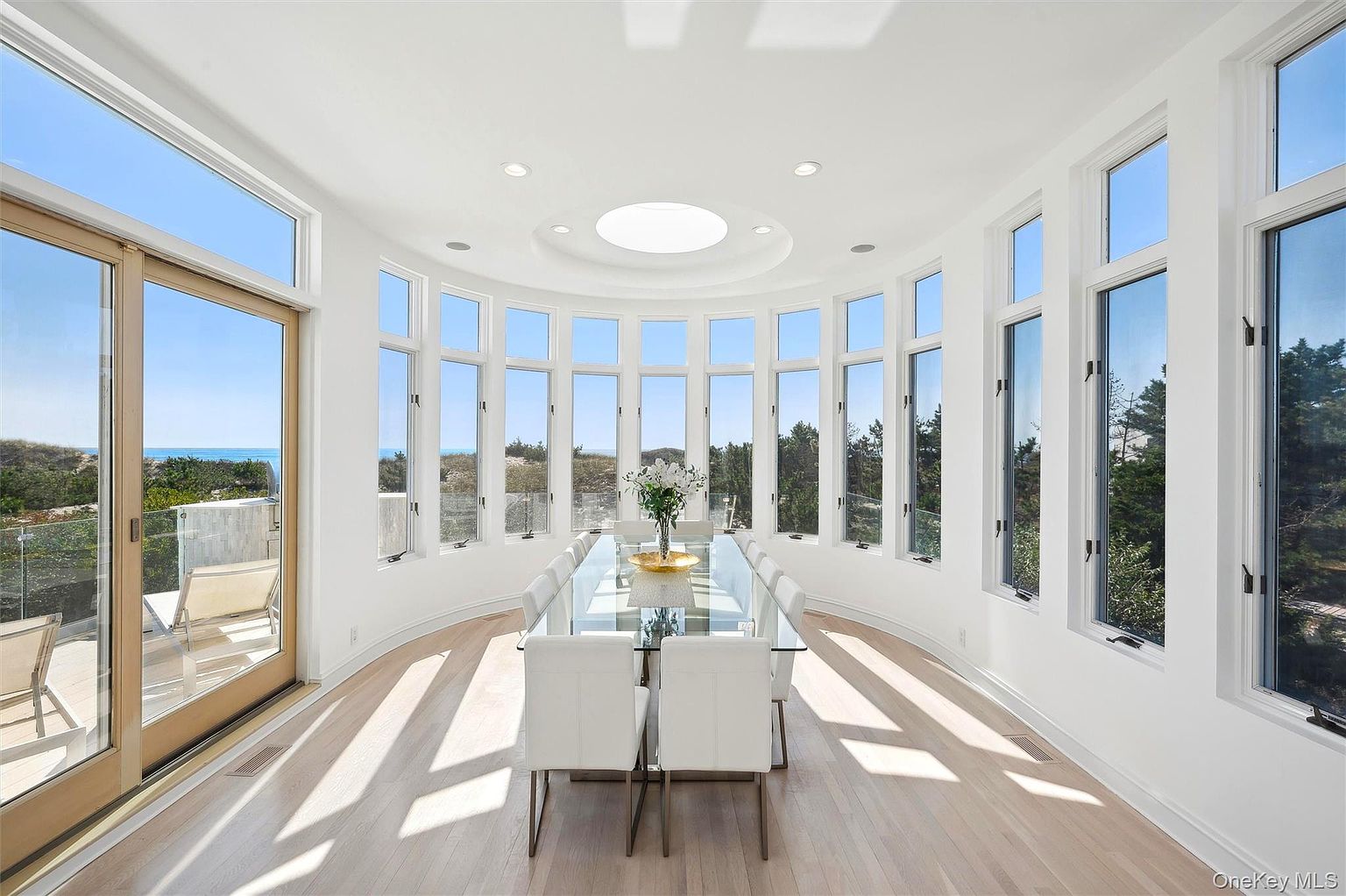 This is a bright and airy dining room featuring a long glass table surrounded by white chairs. The room is enclosed by a curved wall of windows offering panoramic views of the outdoors. A circular skylight provides additional natural light, enhancing the room's spacious and elegant feel.