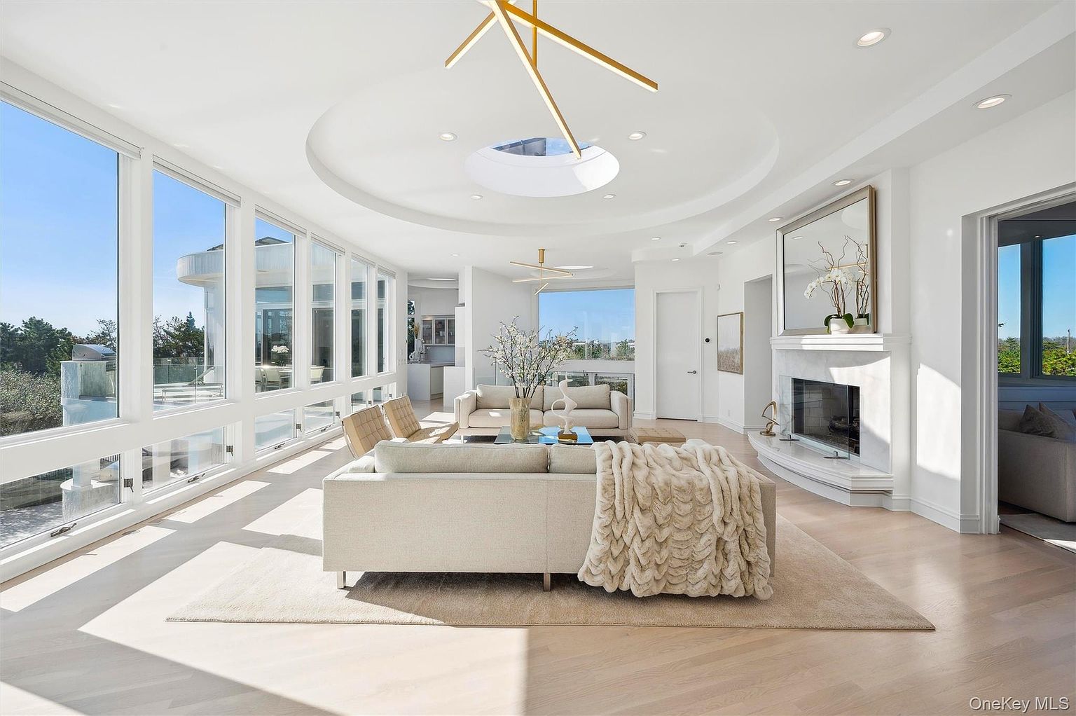 This is a bright and airy living room featuring floor-to-ceiling windows that offer expansive views. The room is decorated in neutral tones with modern furniture, including a large sofa with a cozy throw blanket. A fireplace with a decorative mirror above it adds a touch of elegance, and the overall impression is one of luxury and comfort.