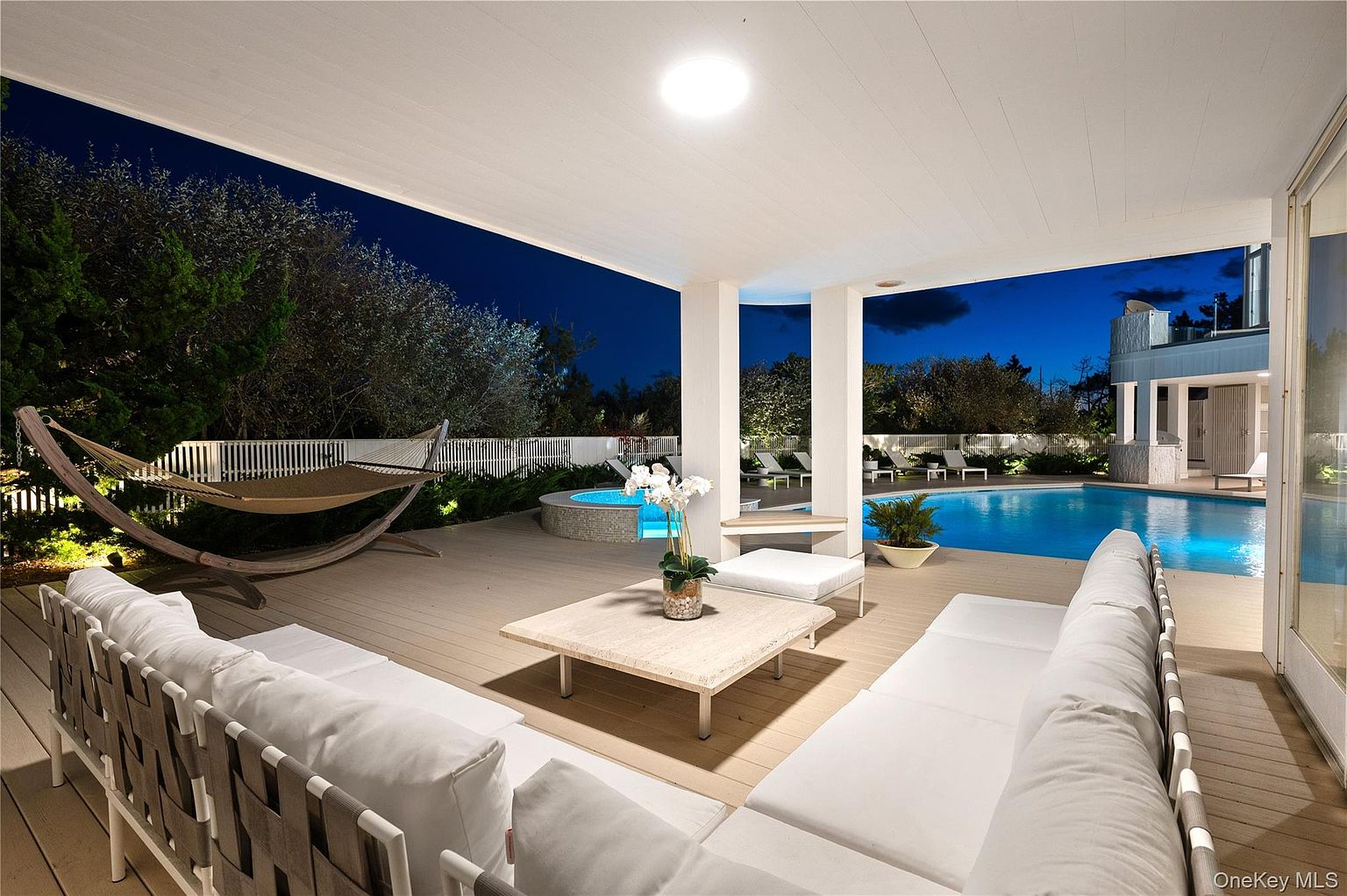 This image showcases a luxurious outdoor patio area at night, featuring comfortable white lounge seating, a hammock, and a modern coffee table with floral decor. A swimming pool and spa are visible in the background, illuminated with blue lighting, creating a serene and inviting atmosphere. The space is well-lit and designed for relaxation and entertainment.