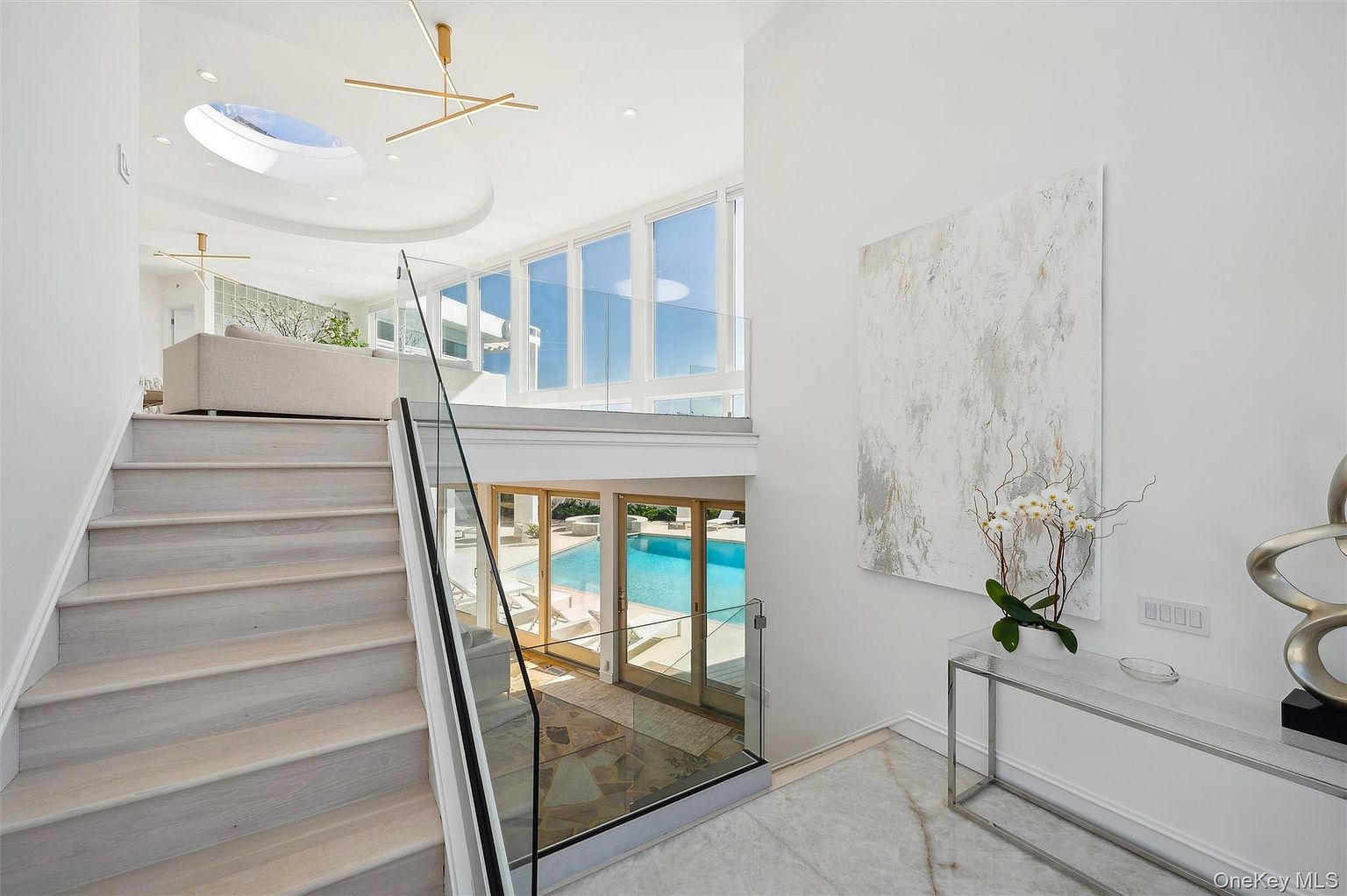 This interior shot showcases a bright and modern hallway with a staircase leading to an upper level. The staircase features light wood steps and a glass railing, offering a clear view of the pool area through large glass doors. A contemporary painting and a sleek console table with floral arrangement add to the sophisticated ambiance.
