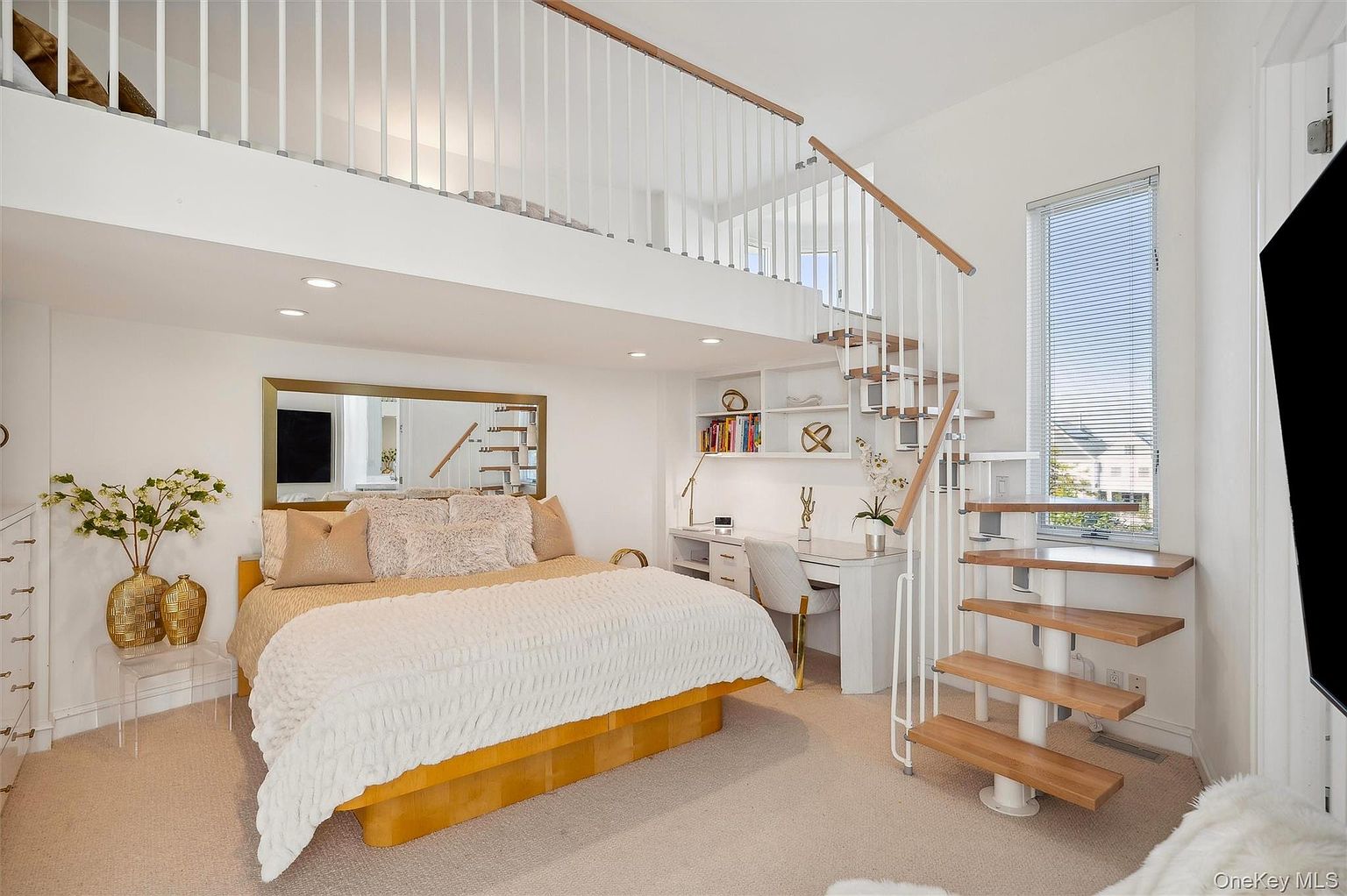This is a bright and airy primary bedroom featuring a unique loft space accessible by a spiral staircase. The room includes a bed with neutral-toned bedding, a large mirror, and a built-in desk area with shelving. The overall design is modern and maximizes vertical space, creating a functional and stylish retreat.