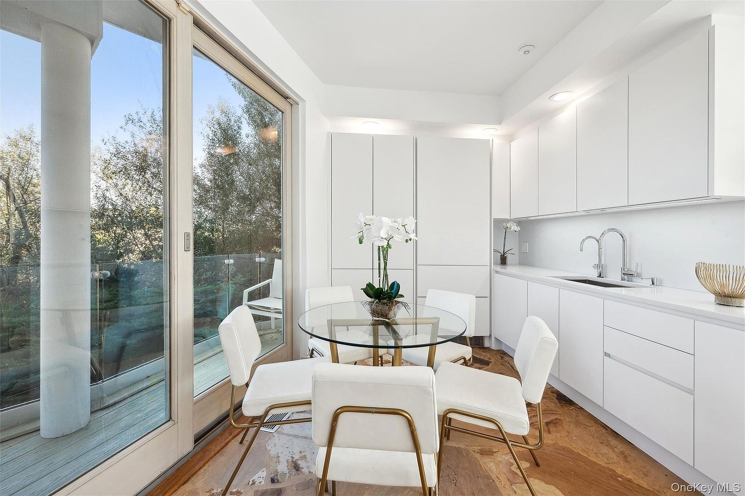 This is a bright and modern kitchen featuring sleek white cabinetry and a unique round glass dining table with gold-toned chairs. The room is well-lit, with natural light streaming in from a large sliding glass door that leads to a balcony, and recessed lighting illuminating the countertops and cabinets. The flooring appears to be a patterned stone or tile, adding a touch of elegance to the space.