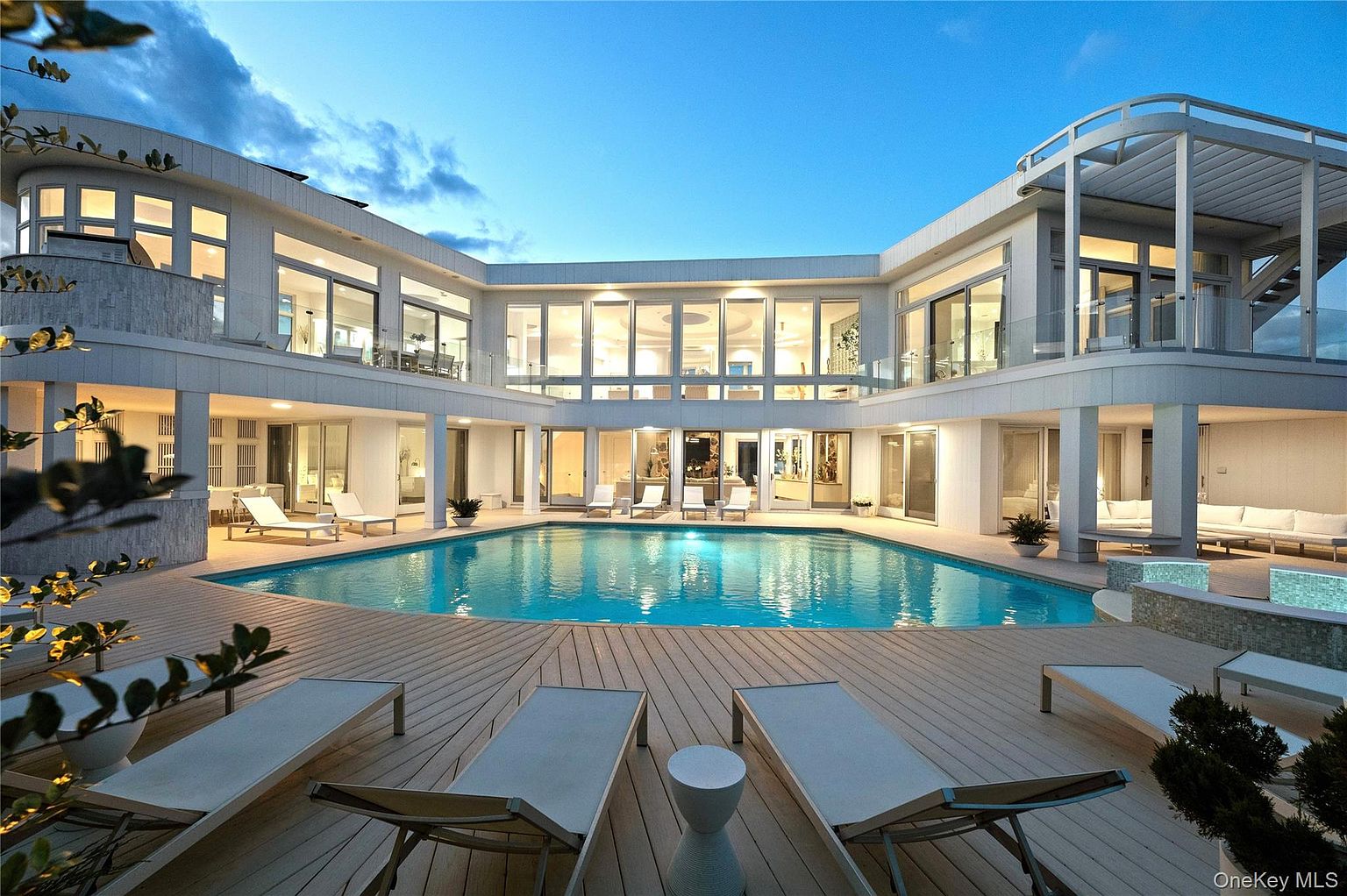 This image showcases a luxurious backyard featuring a large, inviting pool surrounded by a wooden deck. Several lounge chairs are arranged around the pool, creating a relaxing atmosphere. The modern architectural design of the house, with its expansive windows and clean lines, complements the outdoor space, suggesting a high-end property.