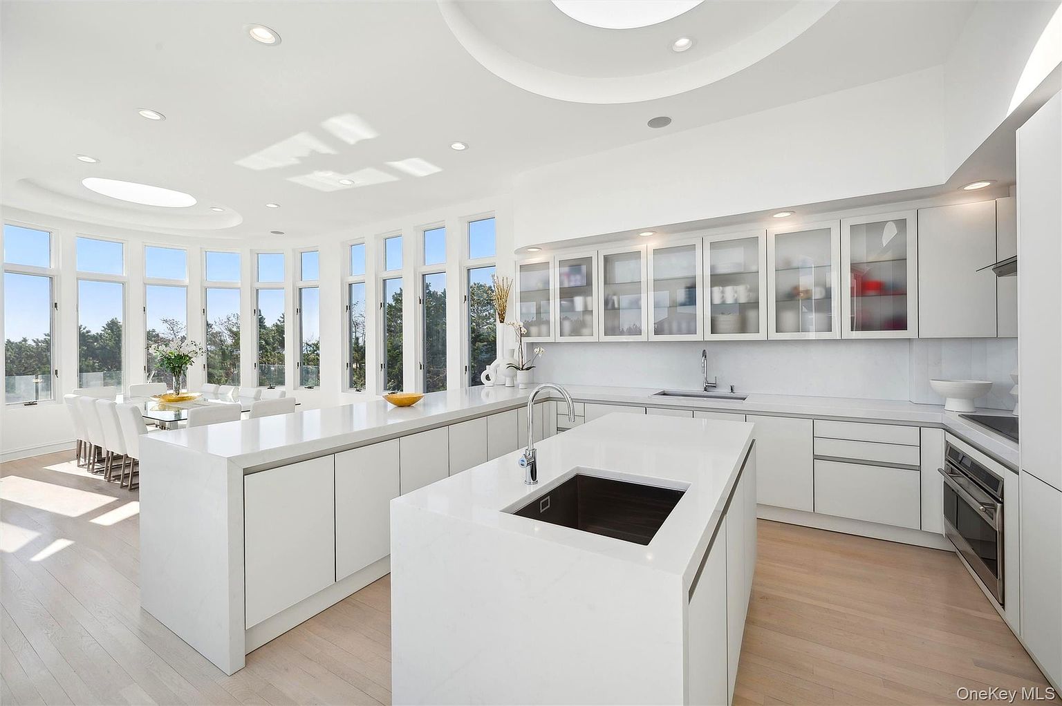 This is a bright and modern kitchen featuring two large islands with white countertops and cabinetry. The kitchen is well-lit with natural light from the surrounding windows and skylights. Stainless steel appliances are integrated into the design, and glass-front cabinets display dishware and other items.
