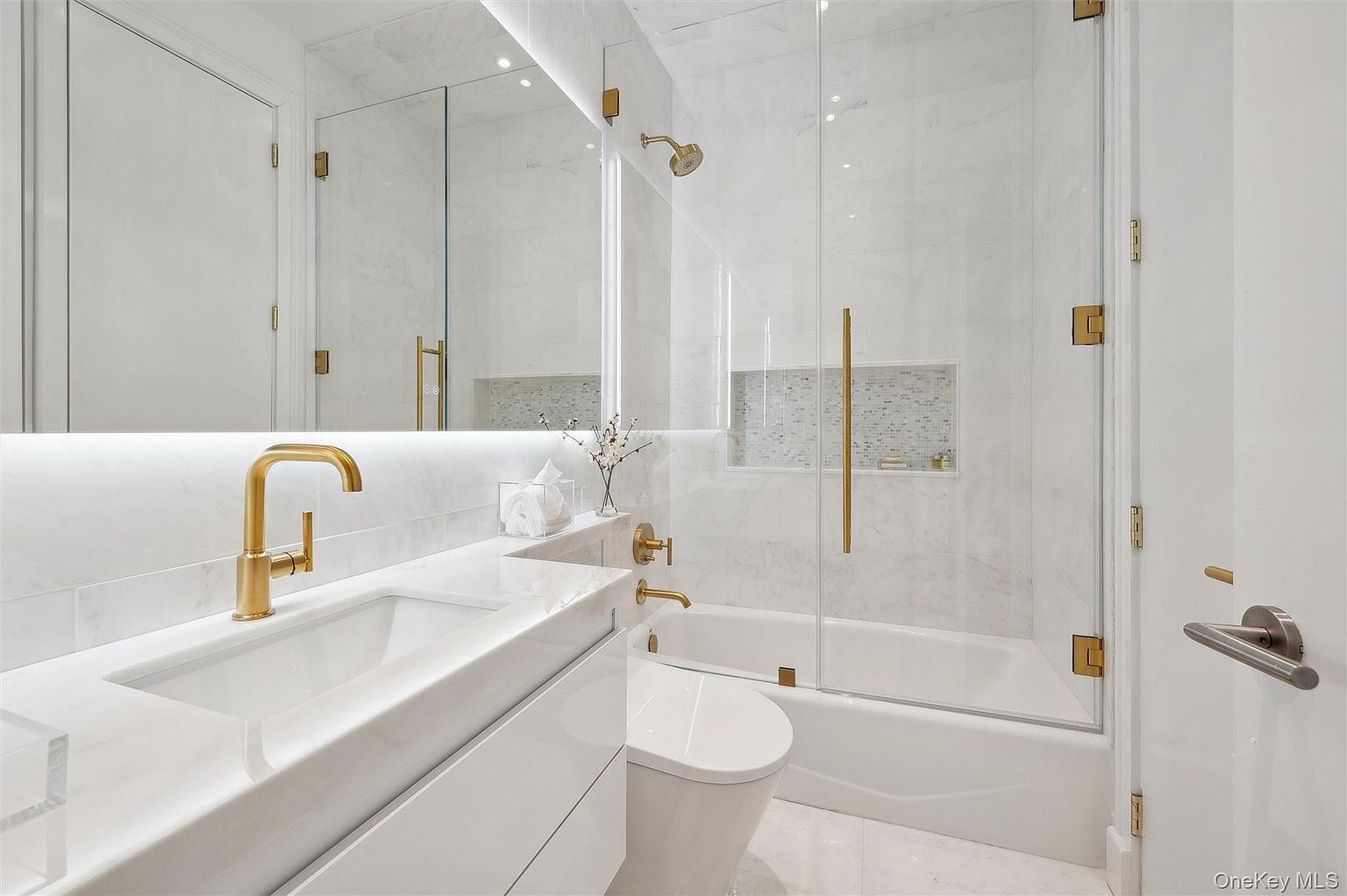 This is a primary bathroom featuring a modern design with white marble walls and countertops, complemented by gold fixtures. A glass-enclosed shower/tub combination is visible, along with a toilet and a sleek vanity with a rectangular sink. The overall impression is luxurious and clean, highlighting the high-end finishes.