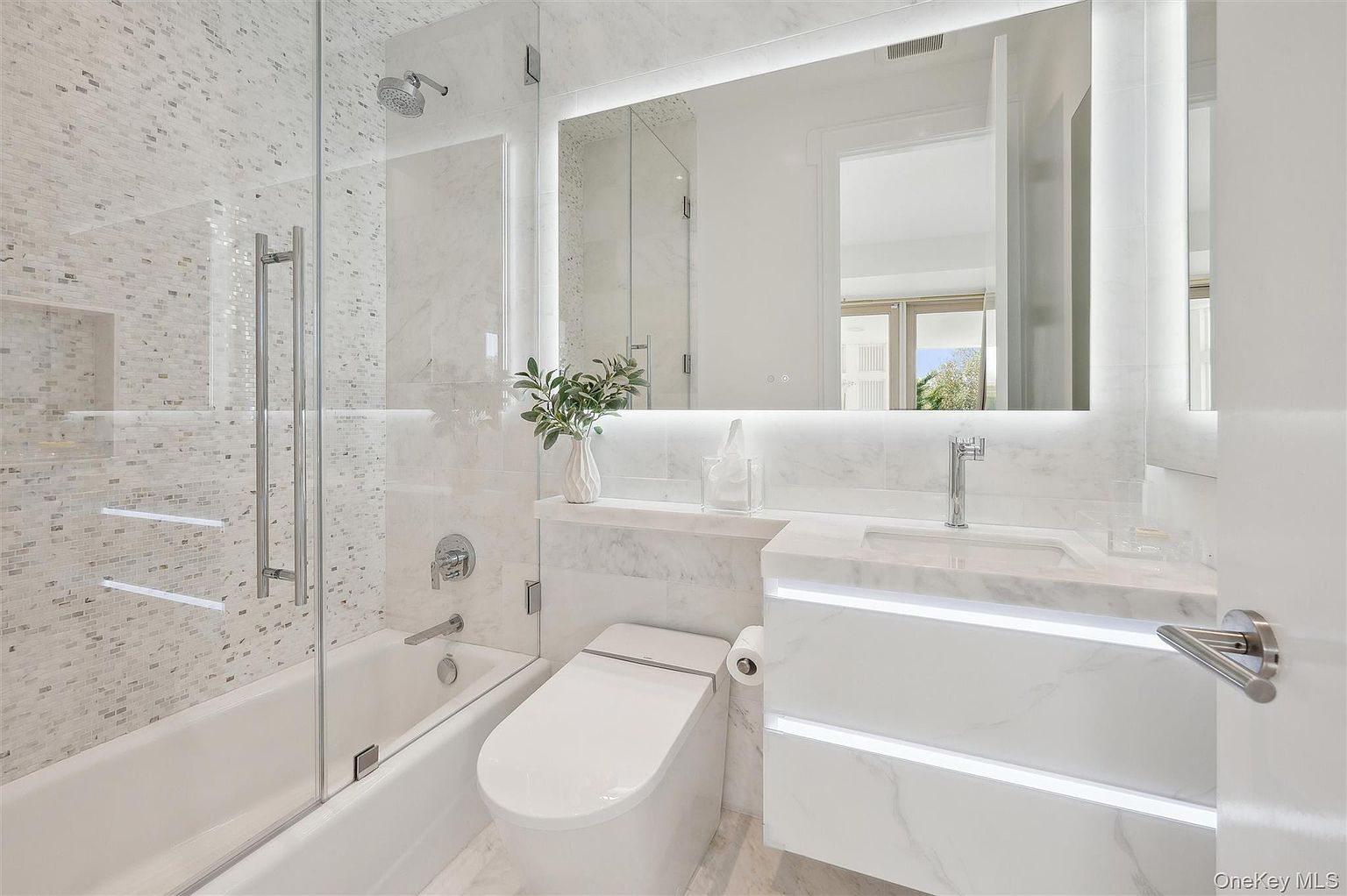 This is a well-lit primary bathroom featuring a bathtub with a glass enclosure, a modern toilet, and a vanity with integrated lighting. The walls and floors are covered in marble, creating a luxurious and clean aesthetic. The large mirror reflects the space, enhancing the sense of openness.