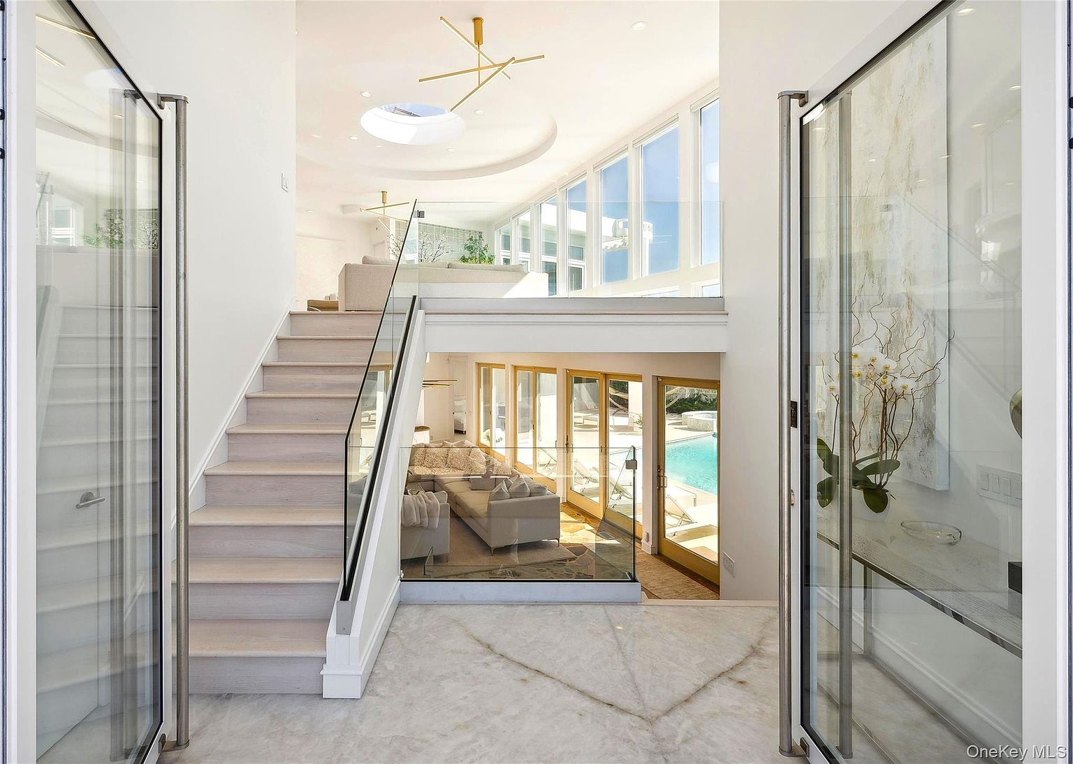 This interior shot showcases a modern home's grand entrance with a focus on the hallway and stairs. The staircase features light wood steps and a glass railing, leading to an upper level with a living area. Below, a glimpse of another living space with a pool view is visible through glass panels, creating a sense of openness and luxury.