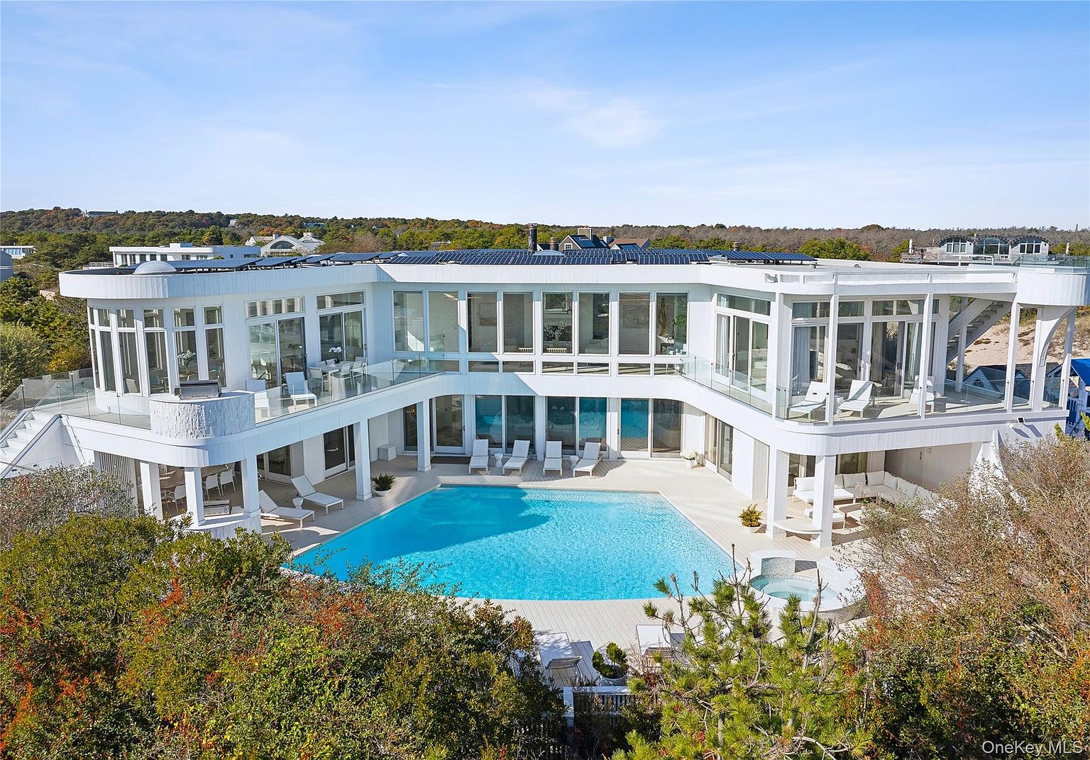 This aerial view showcases a modern, luxurious home with a large swimming pool and spa. The house features expansive glass windows and multiple balconies, offering stunning views. The surrounding landscape includes lush greenery, providing privacy and a serene atmosphere.