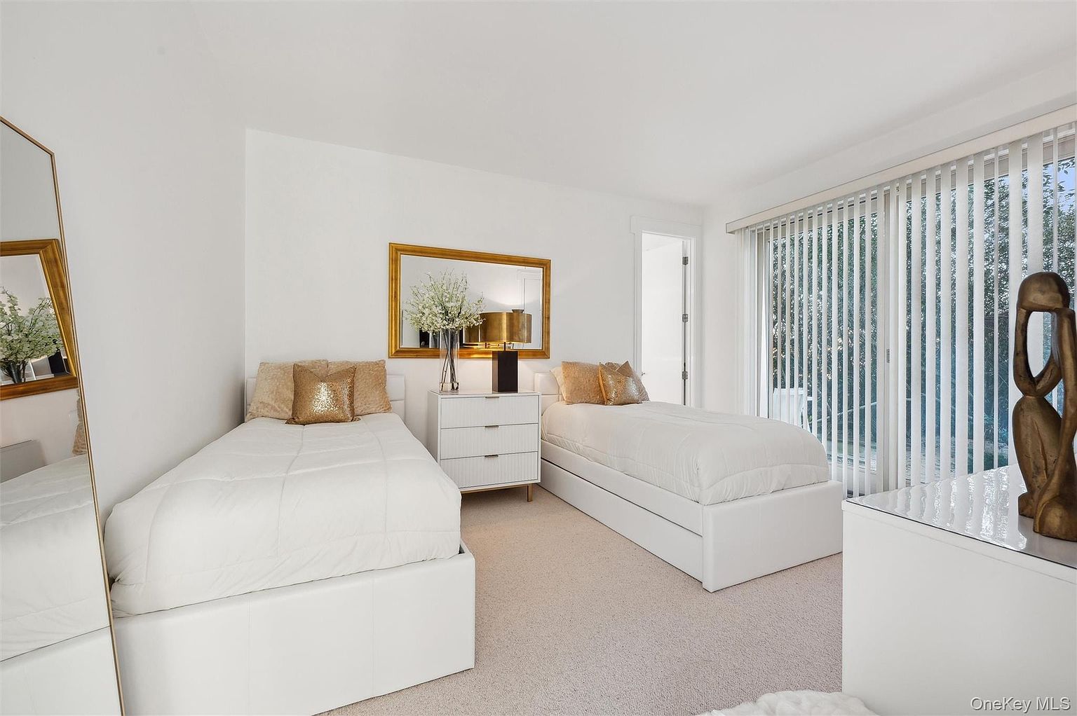 This is a bright and airy guest bedroom featuring two twin beds with white bedding and gold accent pillows. A gold-framed mirror hangs above a white dresser, adding a touch of elegance. Natural light floods the room through large windows, creating a welcoming and comfortable space.