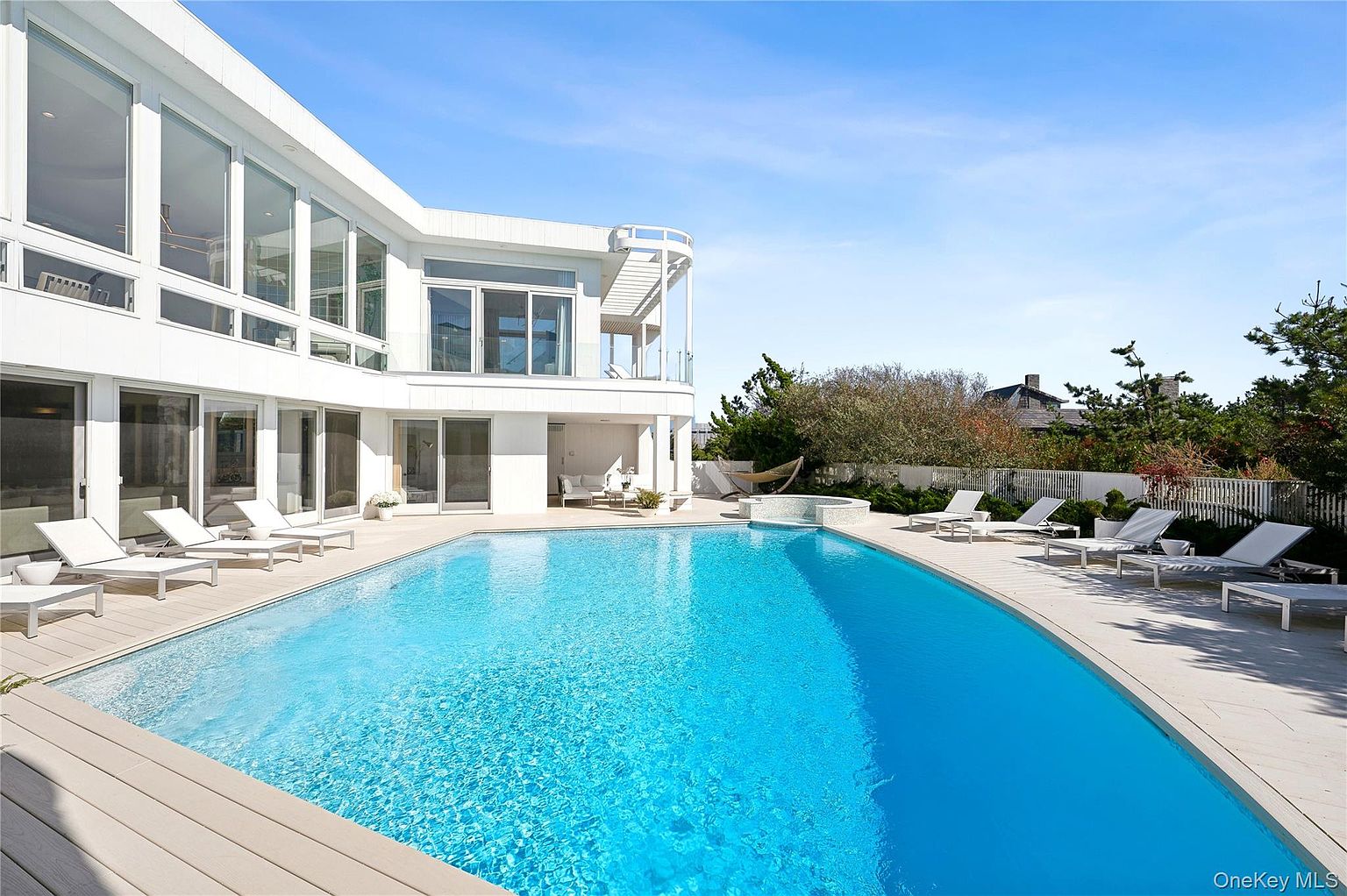 This image showcases a luxurious backyard featuring a large, curved swimming pool with crystal-clear blue water. The pool is surrounded by a light-colored deck with multiple lounge chairs, perfect for relaxation. The modern white house with large windows overlooks the pool area, creating a serene and inviting atmosphere.