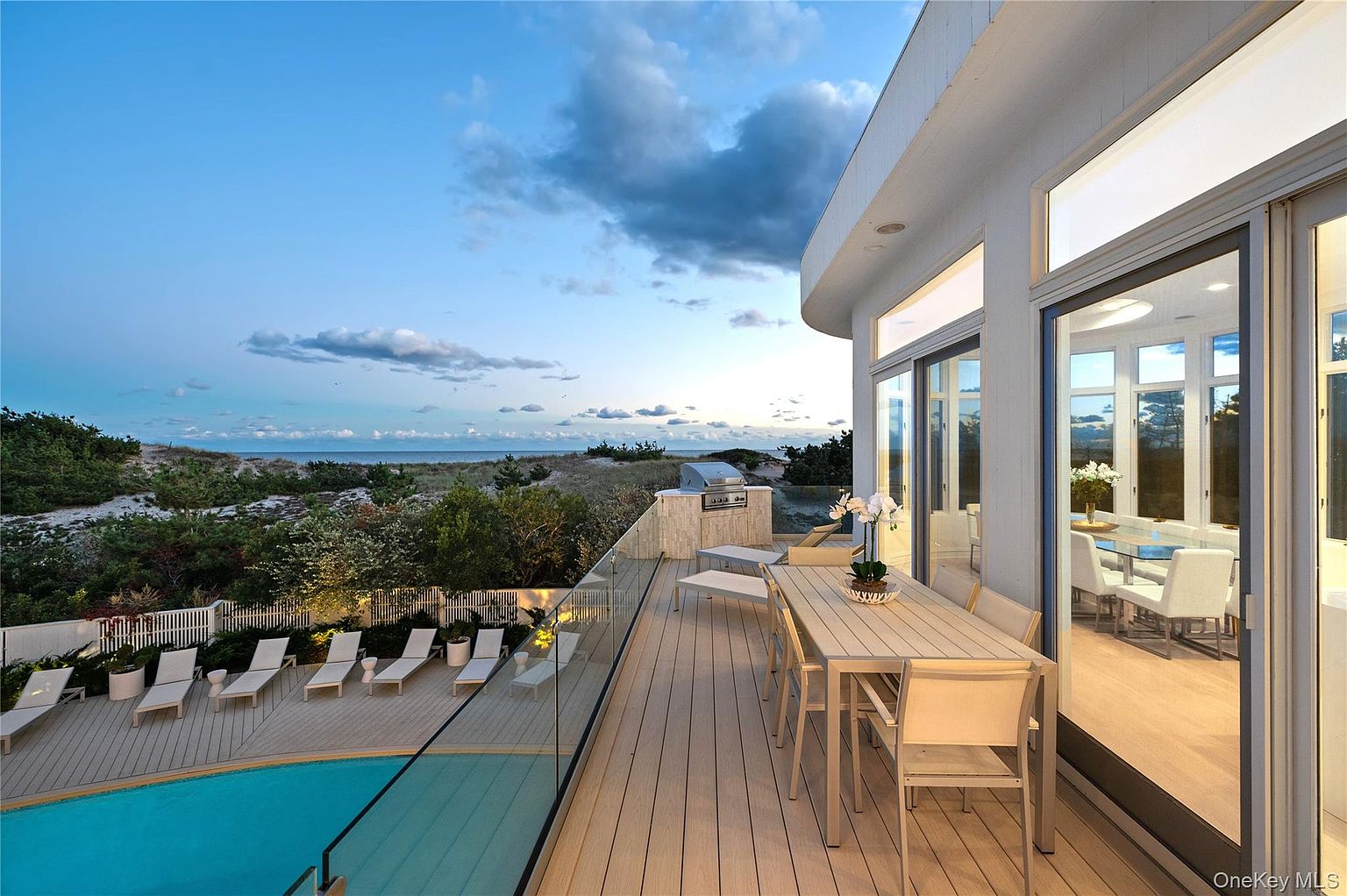 This image showcases a luxurious outdoor living space with a modern aesthetic. The spacious deck features a dining area, lounge chairs, and a built-in grill, all overlooking a pool and the ocean. Large sliding glass doors seamlessly connect the indoor and outdoor spaces, enhancing the property's appeal for entertaining and relaxation.