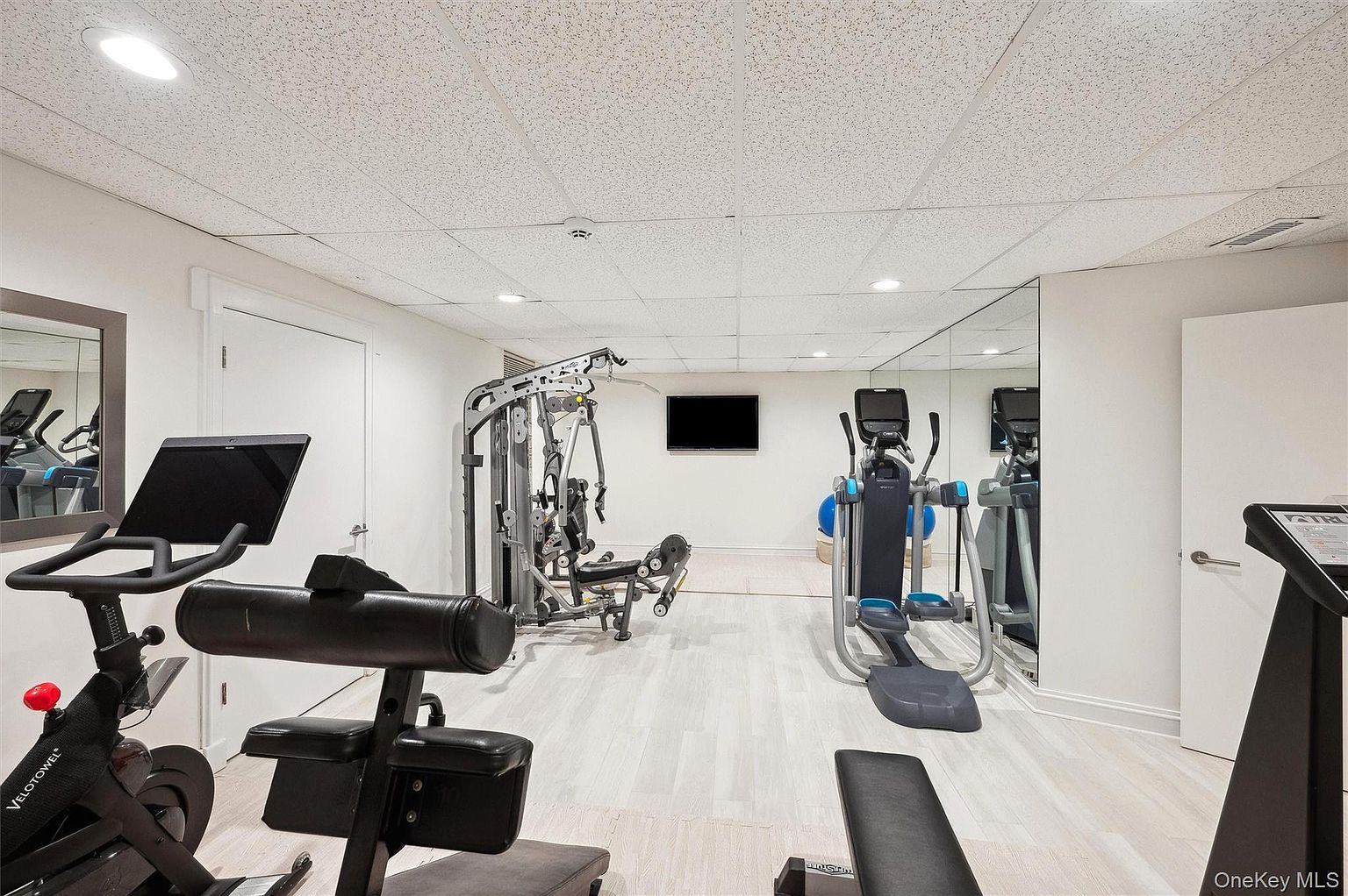 This is an interior shot of a home gym. The room features various exercise machines, including a stationary bike, a multi-station weight machine, and an elliptical trainer. The walls are painted white, and the floor is a light-colored wood or laminate. A large mirror on one wall reflects the equipment, creating a sense of spaciousness.
