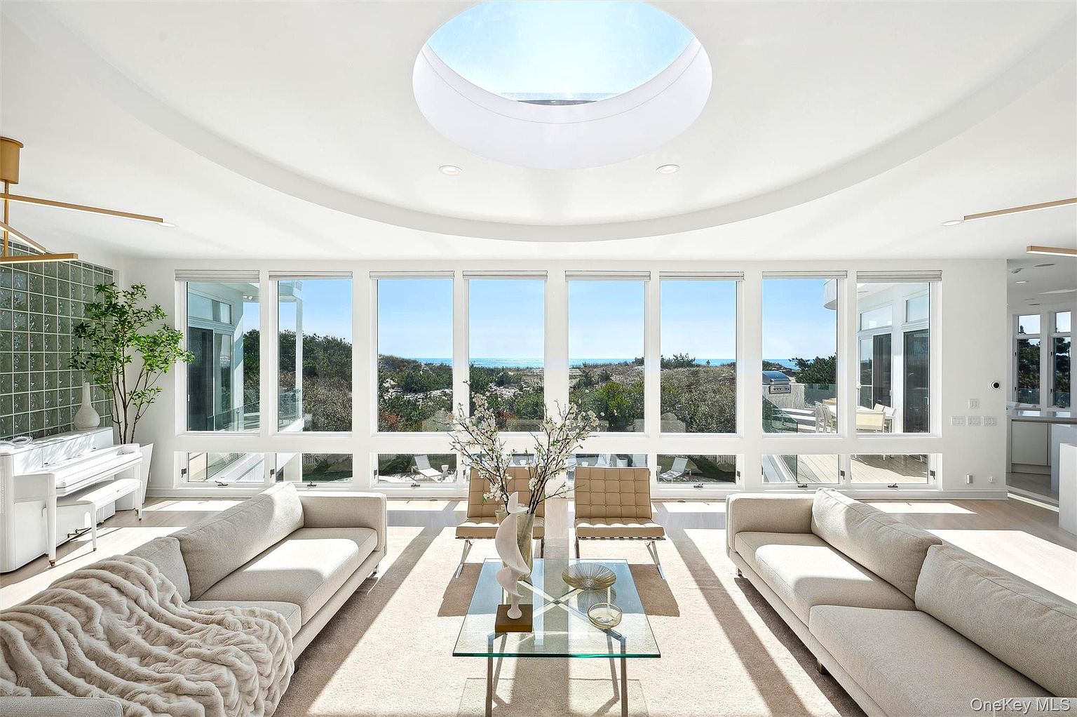 This is a bright and airy living room featuring a wall of windows with views of the ocean and surrounding greenery. The room is furnished with two light-colored sofas, two modern chairs, and a glass coffee table. A skylight adds to the natural light, and the overall style is modern and minimalist, creating a serene and inviting atmosphere.