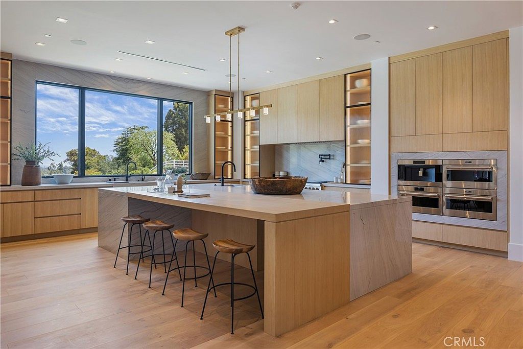 This sophisticated, modern kitchen features a massive waterfall-edge island with light wood cabinetry and matching bar stools, creating a warm and inviting atmosphere. The space is equipped with high-end integrated appliances, sleek light wood cabinetry, and a large window that floods the room with natural light while offering a scenic outdoor view. The minimalist design, combined with the elegant stone backsplash and warm wood flooring, creates a luxurious and functional culinary environment.