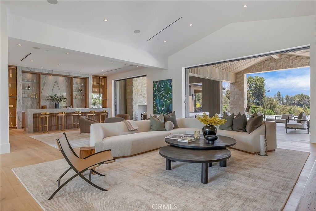 This expansive, open-concept living room features a sophisticated, modern aesthetic with high vaulted ceilings and seamless indoor-outdoor flow through large sliding glass doors. The space is anchored by a plush, curved sectional sofa and a layered circular coffee table, complemented by a sleek bar area with marble accents in the background. The neutral color palette and natural wood flooring create an airy, luxurious atmosphere perfect for high-end entertaining.