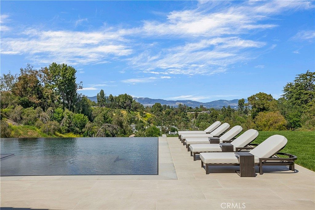 This stunning infinity-edge pool overlooks a lush, rolling hillside landscape under a bright, clear blue sky. A row of sleek, modern lounge chairs is neatly arranged on the expansive stone patio, creating a luxurious and serene outdoor retreat. The perspective captures the seamless transition between the pool's water and the distant mountain horizon, emphasizing the property's high-end, resort-like atmosphere.