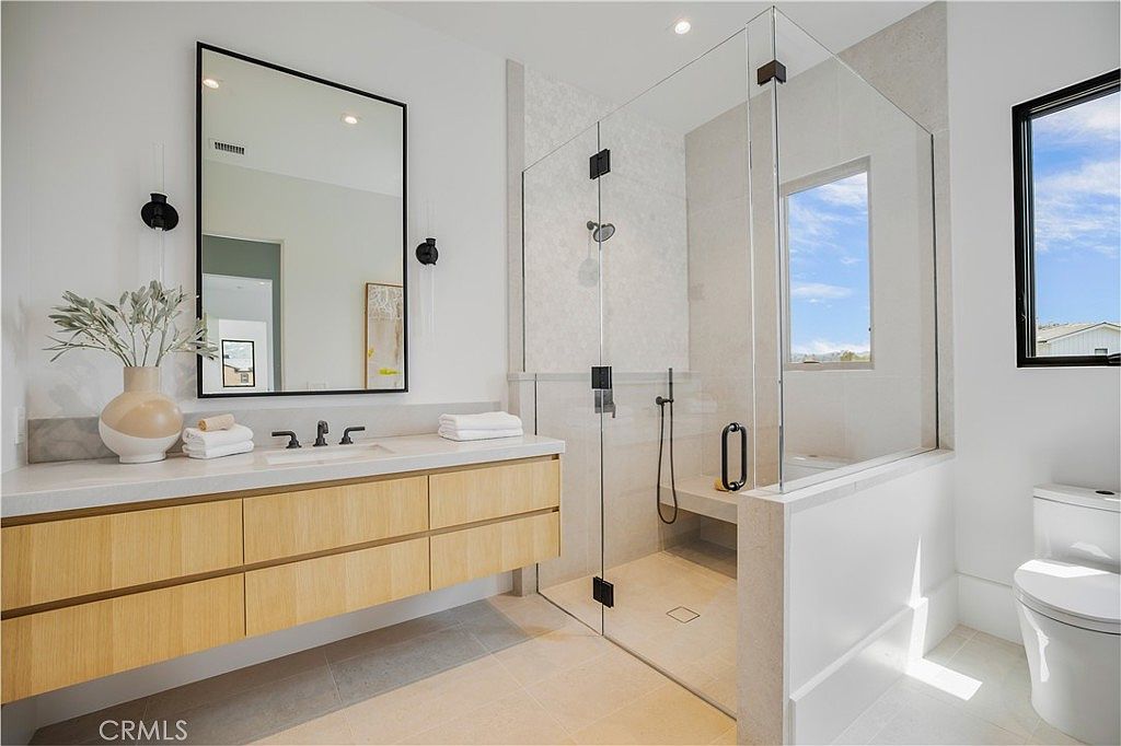 This modern primary bathroom features a sleek floating vanity with light wood cabinetry and a white countertop, paired with a large rectangular mirror. A spacious walk-in shower with a glass enclosure, a built-in bench, and light-toned tile work serves as the focal point. The room is bright and airy, accented by a minimalist toilet and clean, contemporary finishes that create a spa-like atmosphere.