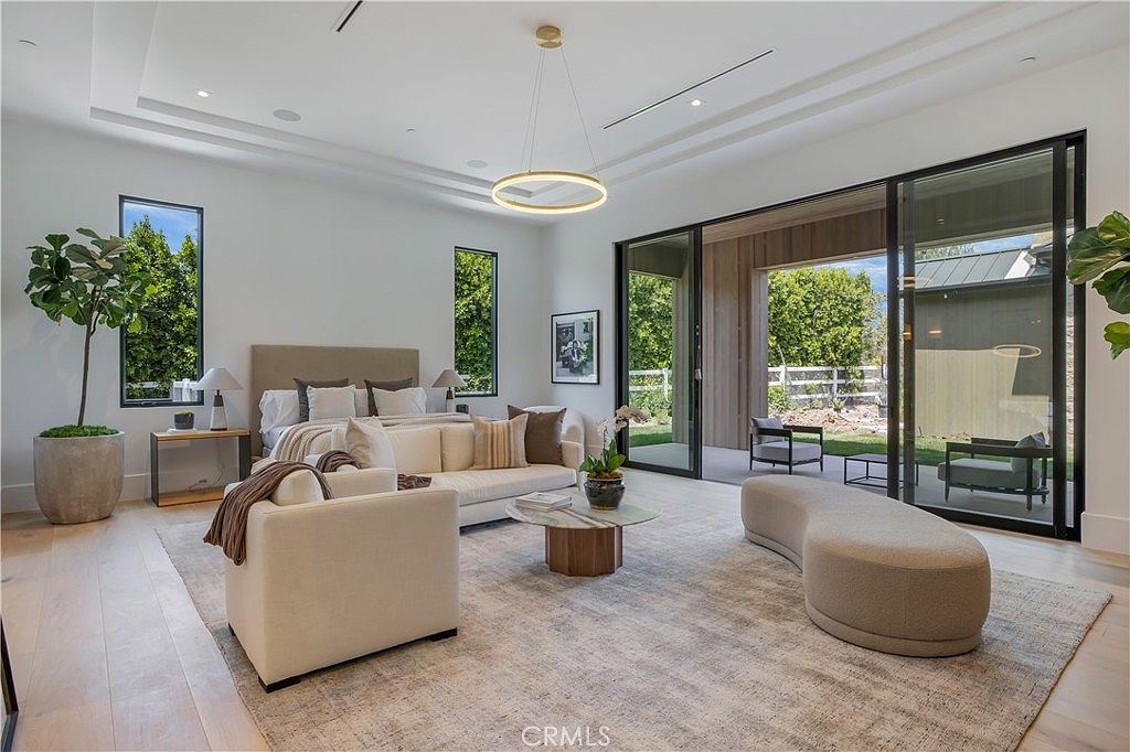 This spacious primary bedroom features a contemporary design with a neutral color palette, a plush king-sized bed, and a comfortable seating area. Large floor-to-ceiling glass sliding doors open to a private patio, seamlessly blending indoor and outdoor living spaces. The room is accented by a modern circular chandelier, a large area rug, and tasteful decor, creating an atmosphere of sophisticated luxury.