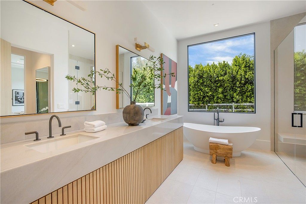 This luxurious primary bathroom features a sleek, modern design with a double vanity topped by a light-veined stone countertop and a slatted wood base. A freestanding soaking tub is positioned elegantly beneath a large window that offers a serene view of lush greenery, creating a spa-like atmosphere. The space is bright and airy, characterized by clean lines, minimalist fixtures, and a sophisticated, neutral color palette.