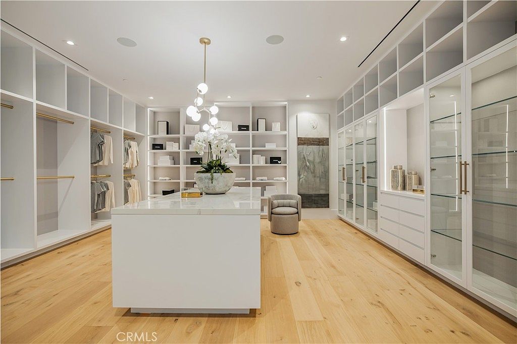 This luxurious, expansive walk-in closet features custom white cabinetry with both open shelving and glass-fronted doors, providing ample storage and display space. A large, marble-topped center island serves as a focal point, complemented by a modern, multi-globe pendant light fixture and light hardwood flooring. The room is bright and airy, offering a sophisticated and organized aesthetic perfect for a high-end primary suite.