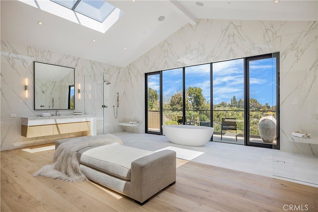 This luxurious primary bathroom features a sleek, modern design with floor-to-ceiling marble walls and light wood flooring. The space is anchored by a freestanding soaking tub positioned in front of expansive glass doors that offer a scenic view, complemented by a minimalist vanity and a plush upholstered bench in the foreground. A large skylight floods the room with natural light, creating an airy and sophisticated spa-like atmosphere.