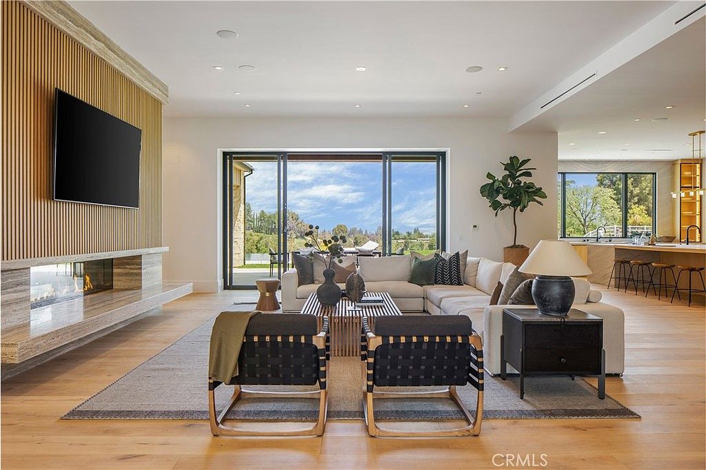 This spacious, open-concept living room features a modern aesthetic with light hardwood flooring, a large sectional sofa, and two stylish accent chairs. A striking floor-to-ceiling slatted wood wall serves as a focal point, housing a sleek fireplace and a wall-mounted television, while expansive sliding glass doors provide a seamless transition to the outdoor space. The room is bathed in natural light, creating an airy and sophisticated atmosphere perfect for entertaining.
