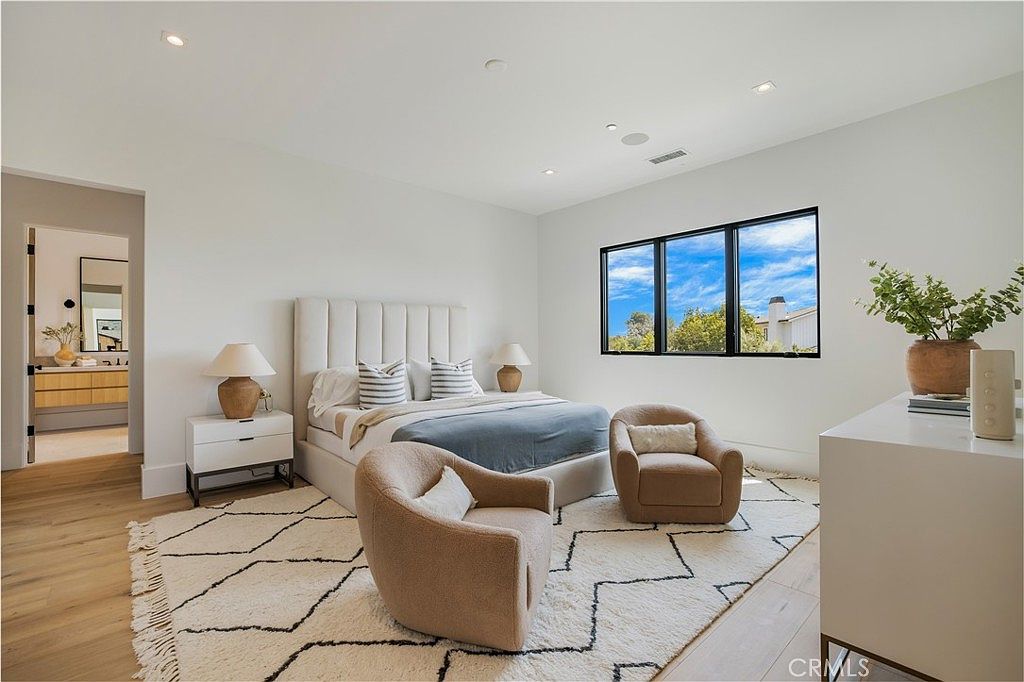 This spacious primary bedroom features a neutral color palette, a plush upholstered bed, and two comfortable accent chairs arranged on a textured area rug. Large black-framed windows allow for ample natural light, while a clean, minimalist aesthetic creates a serene and inviting atmosphere. The room also includes a glimpse into an adjoining bathroom, highlighting the open and airy layout of the home.