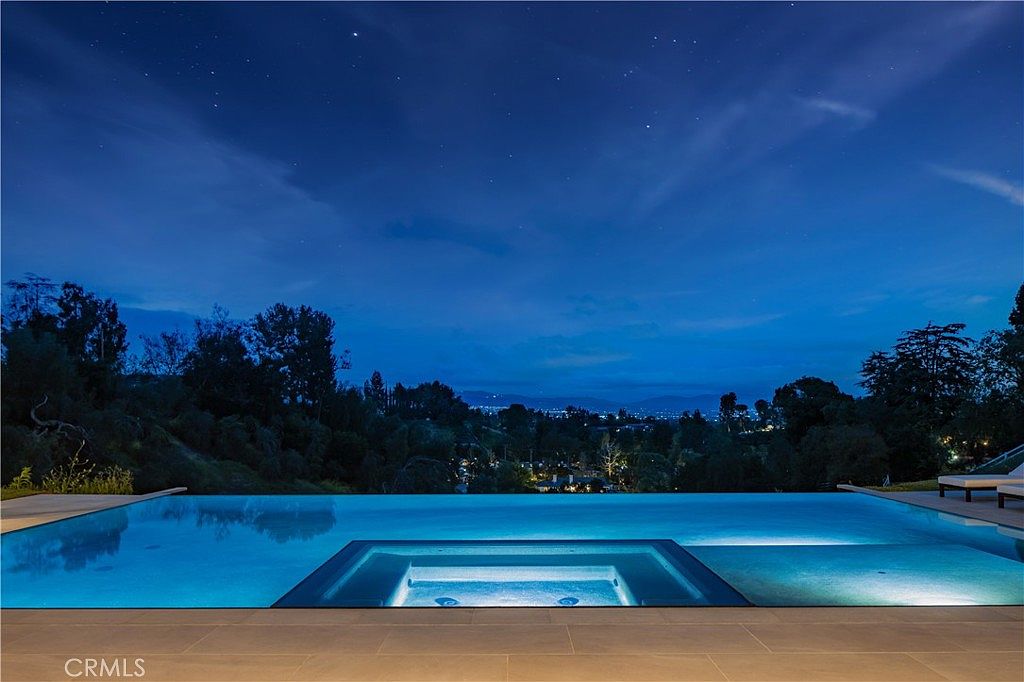 This stunning infinity-edge pool overlooks a vast, twilight landscape, creating a serene and luxurious atmosphere. The pool features a built-in spa section and is framed by a clean, modern stone deck that seamlessly blends into the horizon. The deep blue evening sky and distant city lights provide a dramatic and sophisticated backdrop for this high-end outdoor living space.