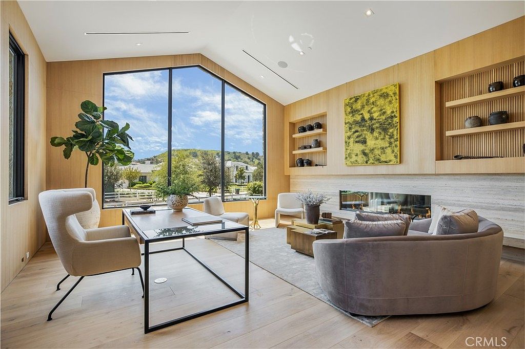 This sophisticated home office features a contemporary design with light wood paneling, a sleek glass-top desk, and a curved velvet sofa. Large floor-to-ceiling windows flood the space with natural light and offer a scenic view of the outdoors, while a built-in fireplace and shelving add warmth and character. The room exudes a professional yet comfortable atmosphere, perfect for focused work or relaxed meetings.
