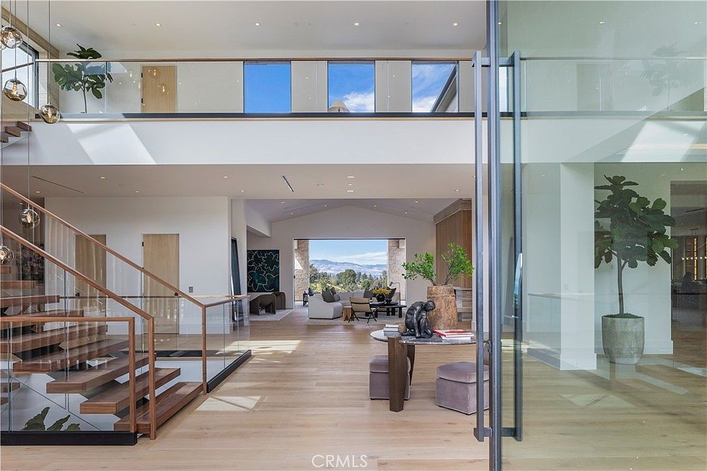 This grand, open-concept entryway showcases a striking floating staircase with glass railings and warm wood treads, leading to an expansive upper landing. The space flows seamlessly into a bright living area with floor-to-ceiling glass doors that frame a scenic mountain view, emphasizing a luxurious, modern aesthetic. High ceilings, light hardwood flooring, and minimalist decor create an airy, sophisticated atmosphere perfect for high-end real estate.